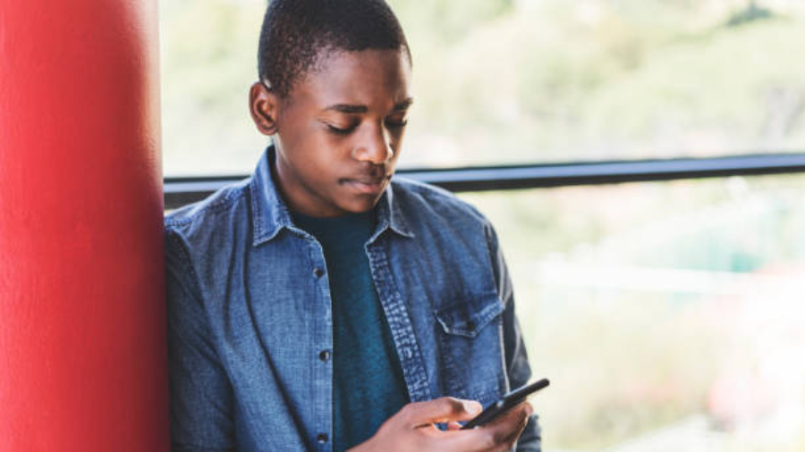 Teenage boy using smart phone against window. Male high school student is leaning on red column. He is wearing denim shirt in school building.